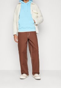 Giacca in pile crema, felpa con cappuccio azzurro chiaro, pantaloni marroni con cuciture bianche e sneakers crema. Texture morbide e vestibilità rilassata.