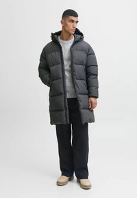 Abrigo acolchado gris oscuro con capucha, diseño acolchado y cremallera frontal. Combinado con un suéter gris claro y pantalones negros, llevado con zapatos beige.