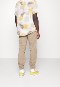 Kortärmad skjorta med ett lätt blommönster i gult och brunt, kombinerad med beiga avsmalnande byxor och sneakers med gula accenter.