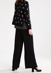Cardigan noir à motif floral, superposé à un haut à pois, associé à un pantalon noir à jambes larges et des talons noirs.