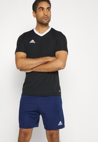 Camiseta deportiva negra con cuello en V blanco y logo de Adidas; combinada con pantalones cortos azul marino con un detalle de logo blanco.
