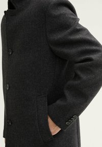 Cappotto herringbone grigio scuro con colletto alto, chiusura con bottoni, tasche laterali e quattro bottoni sulle maniche. Design ispirato all'abbigliamento militare.
