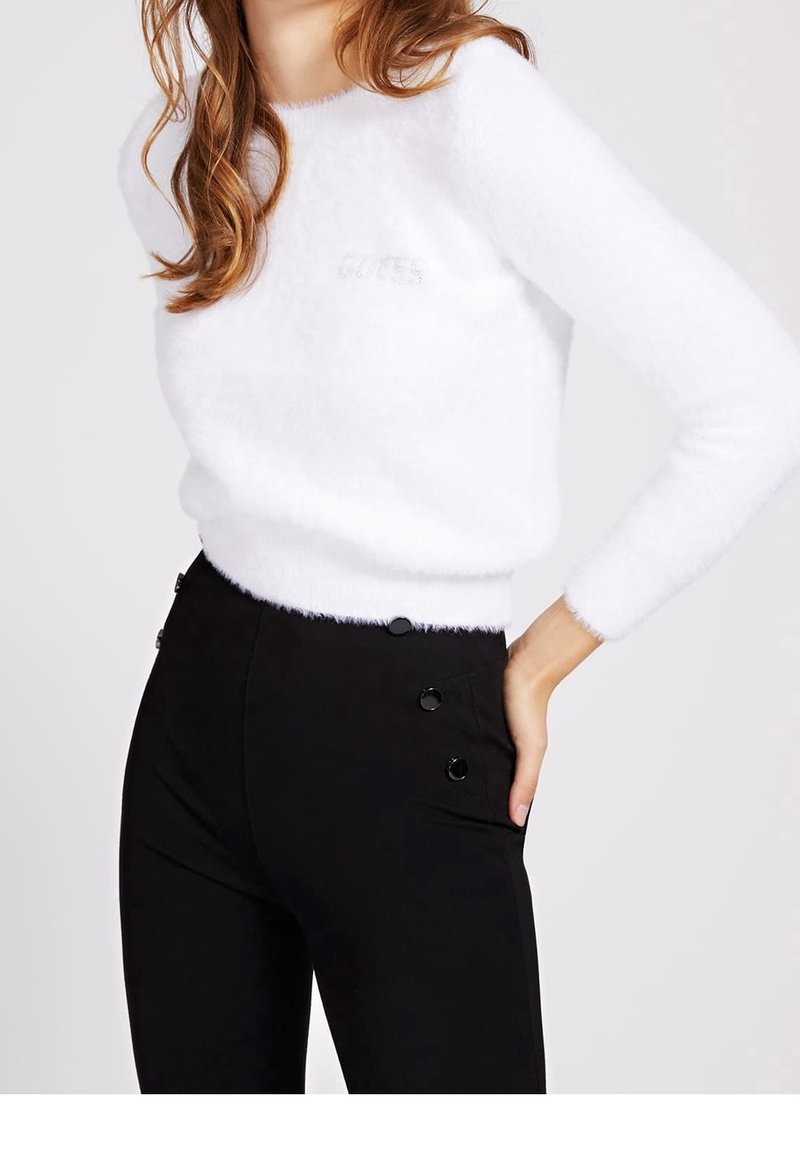 Pull blanc duveteux avec un col rond et le logo "GUESS", associé à un pantalon noir ajusté avec des boutons décoratifs à la taille.