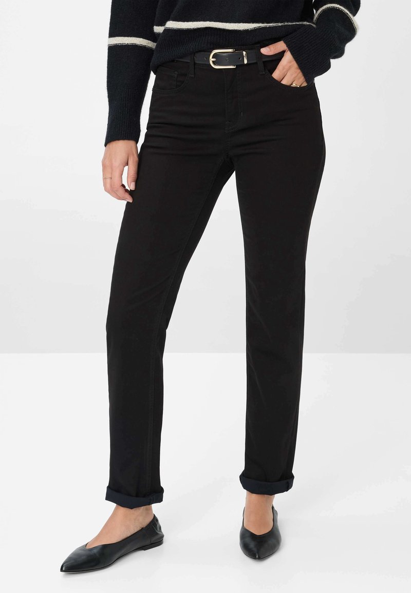 Femme portant un jean noir droit avec des revers, ceinture noire avec boucle dorée, ballerines noires pointues et pull noir à rayures blanches.