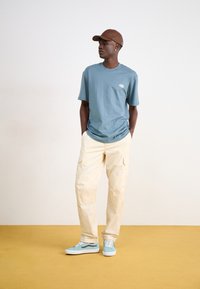 Light blue short-sleeve t-shirt, cream cargo pants, and mint green sneakers. Brown cap and small logo detail on shirt. Smooth texture.
