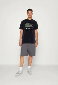 Νεαρός άνδρας στέκεται φορώντας μαύρο τ-shirt Lacoste με το λογότυπο κροκόδειλου, γκρίζες βερμούδες, λευκές κάλτσες και λευκά αθλητικά παπούτσια μπροστά από απλό φόντο.