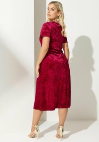 Vestido de terciopelo rojo con mangas cortas, diseño a media longitud y una textura suave. Presenta una cintura ajustada y un drapeado natural. Tacones con tira en el tobillo de color dorado.