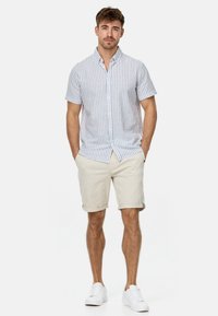 Camisa de manga corta abotonada en rayas verticales azul claro y blanca, combinada con pantalones cortos beige hasta la rodilla y zapatillas blancas.