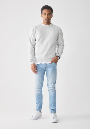Ung man som bär en ljusgrå munkjacka, vita sneakers, ljusblå jeans och en armbandsklocka, står med en hand i fickan mot en vit bakgrund.