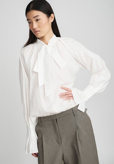 Blouse blanche à manches longues avec un col cravate et des épaules bouffantes, accompagnée de pantalons en houndstooth gris avec des plis et des poches latérales.