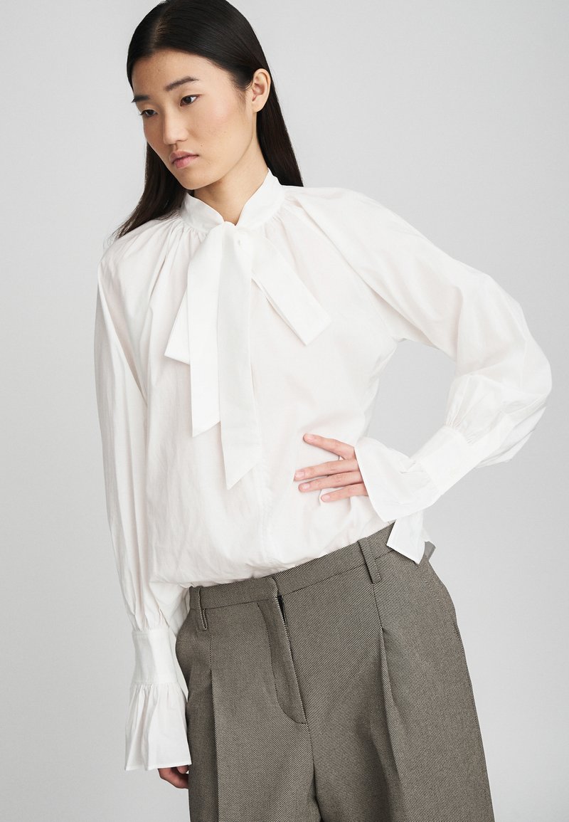 Blusa bianca a maniche lunghe con colletto a fiocco e spalle a sbuffo, abbinata a pantaloni grigi a scacchi houndstooth con pinces e tasche laterali.