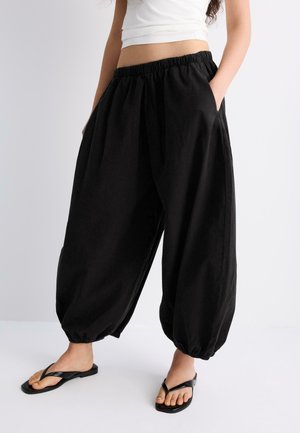 Frau trägt lockere schwarze Haremshosen mit elastischem Bund und Bündchen, weißes kurz geschnittenes Oberteil und schwarze Flip-Flops, steht mit den Händen in den Taschen.