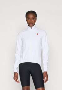 Weiße, leichte Fahrradjacke mit hohem Kragen, Reißverschluss, dehnbaren Bündchen und rotem Logo auf der linken Brust. Tragen mit schwarzen Fahrradhosen.