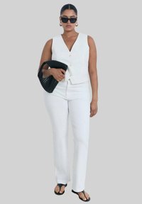 Gilet bianco senza maniche con scollo a V e bottoni, abbinato a pantaloni bianchi. Tiene una borsa nera con texture e indossa occhiali da sole.