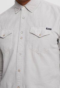 Camicia in corduroy grigio chiaro con colletto a punta, due tasche sul petto e bottoni a pressione in metallo. Presenta un'etichetta con il logo Replay sulla tasca sinistra.