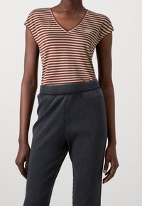 Pasiasty t-shirt z dekoltem w serek w kolorze burgundowym i kremowym, w połączeniu z ciemnoszarymi spodniami z prążkowanej tkaniny. Materiał wydaje się miękki, a fason jest luźny. Obecny jest mały detal z logo.