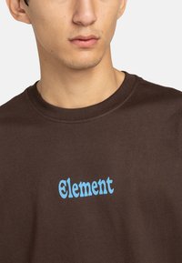Bruine T-shirt van katoen, met een blauw "Element" logo in een speelse lettertype. Ronde hals, korte mouwen en een gladde textuur.