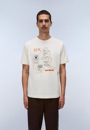 Homme aux cheveux courts portant un t-shirt blanc avec une carte détaillée et un texte japonais imprimé à l'avant, associé à un pantalon marron foncé.