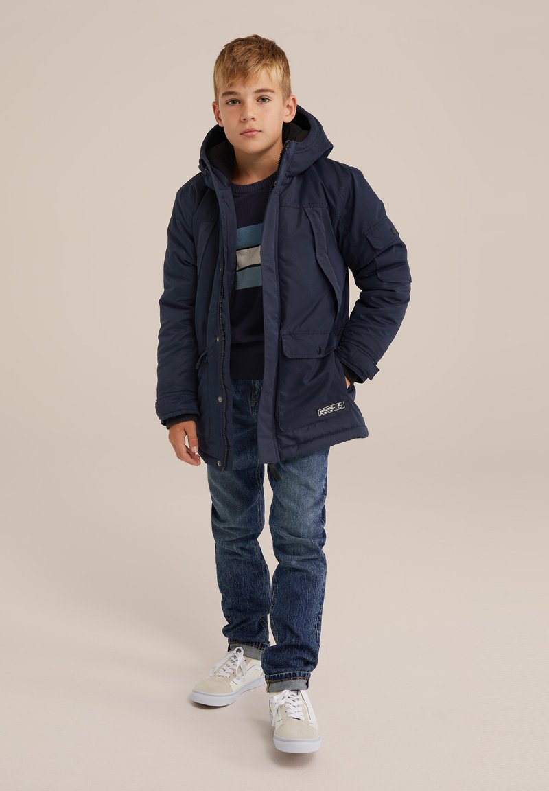 Doudoune navy avec capuche, dotée de poches latérales et d'une fermeture éclair. Portée par-dessus un pull rayé navy et associée à un jean bleu et des baskets blanches.
