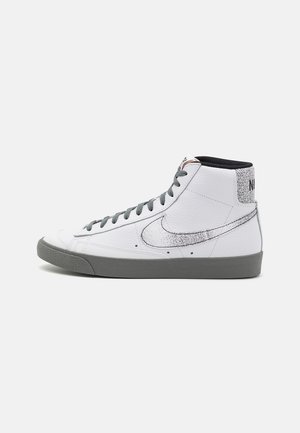Witte hoge sneaker met grijze zool en veters, verweerde zwarte Nike-swoosh en zwart Nike-logo op het hielstuk.