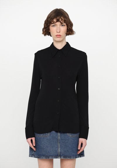 Zwarte blouse met lange mouwen, knoopsluiting, een puntige kraag en een gladde textuur, gecombineerd met een spijkerrok met voorzakken.
