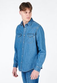 Camicia di jeans azzurra in un blu medio con colletto classico, due tasche frontali, dettagli di doppia cucitura e maniche lunghe.