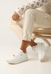 Białe sneakersy z teksturowanymi panelami i złotymi akcentami. Posiadają sznurowany design i gumową podeszwę. Noszone z kremowymi spodniami.