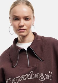 Sudadera de color marrón oscuro con cuello de canalé, media cremallera y texto bordado en blanco. Textura suave con cremallera y diseño de gran tamaño.
