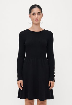 Robe noire côtelée avec des manches longues et un corsage ajusté, s'évasant à la taille. Présente des accents de boutons sur les poignets. Texture lisse.