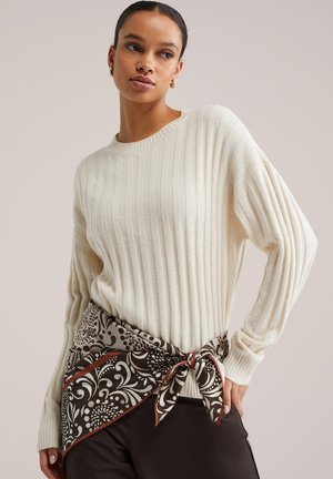 Cremefarbener, gerippter Pullover mit überschnittenen Schultern, kombiniert mit einem gemusterten braun-cremefarbenen Schal, der in der Taille gebunden ist. Glatte Textur und lockere Passform.