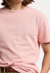 Helles rosa Baumwoll-T-Shirt mit Rundhalsausschnitt und kurzen Ärmeln. Das Shirt hat ein kleines, tonales gesticktes Logo auf der linken Brust. Weicher Stoff.