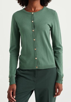 Vrouw draagt een lange mouw groene cardigan met gouden knopen, gecombineerd met donkergroene broek, staand tegen een lichte achtergrond.