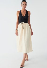 Svart ribbad ärmlös topp med indragen midja, krämfärgad fladdrig midikjol med texturerat mönster och svarta remsandaler.