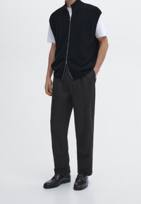 Svart stickad väst med hög krage och dragkedja framtill, bärs över en vit t-shirt. Matchas med lösa svarta byxor och svarta skor.