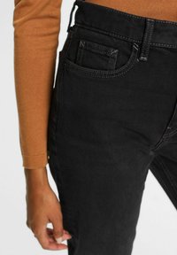 Personne portant un jean en denim noir et un haut marron à manches longues, montrant un gros plan de la poche du jean et du bas du torse.