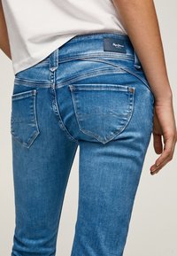 Modré džíny z denimu s obepínajícím střihem, které mají zadní kapsy se špičatým tvarem a koženou nášivku s logem na pase.