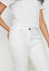 Vaqueros de denim blancos de corte ajustado, con cierre de botón, pasadores para cinturón y dos bolsillos frontales. Textura suave y diseño limpio.