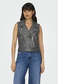 Graue lederne ärmellose Bikerjacke mit großem Kragen, mehreren Reißverschlusstaschen und einem Gürtel in der Taille. Getragen über blauen weiten Jeans.