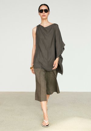 Femme portant un haut asymétrique sans manches foncé, un pantalon large marron raccourci, des sandales, des lunettes de soleil et des bracelets dorés marchant vers l'avant.