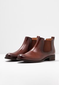 Un par de botas Chelsea de cuero marrón pulido con paneles elásticos en los laterales y tacones bajos apilados sobre una superficie blanca.