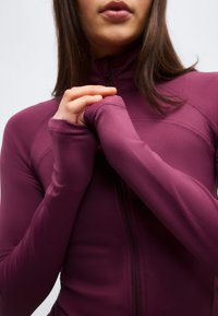 Γυναίκα που φοράει εφαρμοστό, μωβ σακάκι με μακριά μανίκια και τρύπες για τους αντίχειρες, κρατώντας τα χέρια της κοντά στο στήθος της μπροστά σε απλό φωτεινό φόντο.