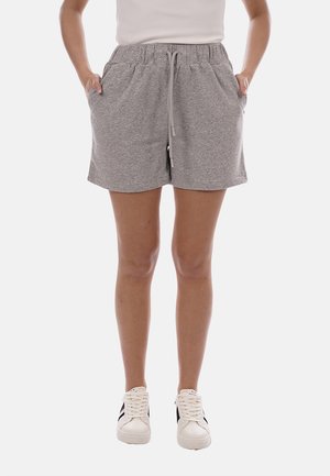 Persona che indossa pantaloncini grigi chiari a fantasia con coulisse, scarpe da ginnastica bianche con strisce nere e un top bianco senza maniche, con le mani nelle tasche.