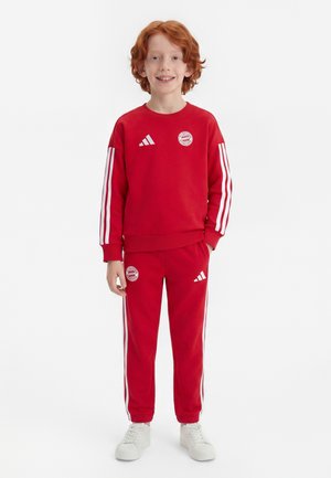 Niño con cabello rojo que lleva un chándal deportivo rojo con rayas blancas y zapatillas blancas, de pie y sonriendo contra un fondo blanco.
