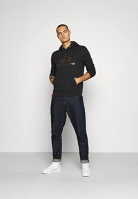 Felpa nera con cappuccio e tasca a marsupio, decorata con un logo in lettere color rame, abbinata a jeans scuri e sneakers alte bianche. Tessuto dalla consistenza liscia.