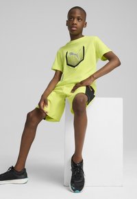Cămașă cu mânecă scurtă de culoare verde lime, cu un design de logo negru, asortată cu pantaloni scurți care au accente laterale negre. Completară cu pantofi sport negri.