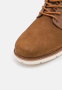 Botín de ante marrón con punta redondeada, suela de goma blanca y cordones beige. Presenta acentos de tejido texturizado en los laterales. Diseño limpio.