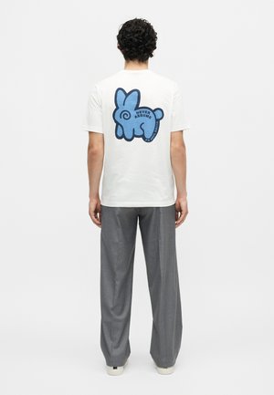 Άνδρας που φοράει λευκό μπλουζάκι με μπλε αφηρημένο σχέδιο λαγού και το κείμενο "Never Assume" στην πλάτη, γκρι παντελόνια, γυρισμένος πλάτη σε λευκό φόντο.