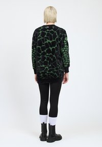 Eine Person mit kurzem blonden Haar steht mit dem Rücken zur Kamera und trägt einen langen schwarz-grünen Musterpullover, schwarze Leggings, weiße Socken und schwarze Stiefel.
