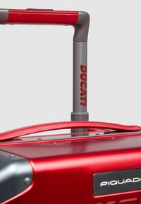Rode harde koffer met een metallic afwerking, voorzien van een grijs telescopisch handvat en het reliëfmerk "DUCATI" op het handvat.