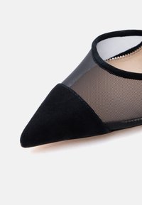 Μαύρο γυναικείο παπούτσι μπαλαρίνα με μυτερή μύτη, διάφανο δίχτυ στο επάνω μέρος και στερεό σουέτ κάλυμμα στη μύτη, σε λευκό φόντο.